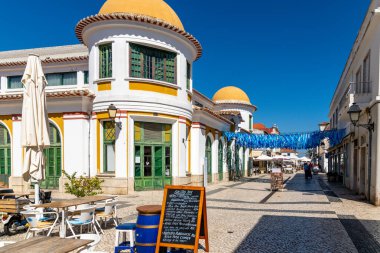 Beyaz-sarı binalı bir pazar meydanı, caddenin üzerinde asılı mavi çelenkler, ön planda şemsiyeli masalar ve sandalyeler.