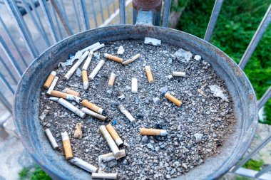 Alghero, Sardinia - İtalya - 02-13-2025: Çakıl ve sigara izmaritli yuvarlak metal bir kül tablası, bazıları yanmış, bazıları ezilmiş. Korkuluklardan önce paslanmış tepsi, çimenler ve arkadaki yürüyüş yolu.