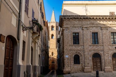 Alghero, Sardinia - İtalya - 02-13-2025: Alghero 'da dar bir sokağın sonunda Gotik bir çan kulesi yükseliyor. Etrafını kumtaşı binalar sarmış. Güneş ışığı, mavi gökyüzünün altında gölgeler bırakır..