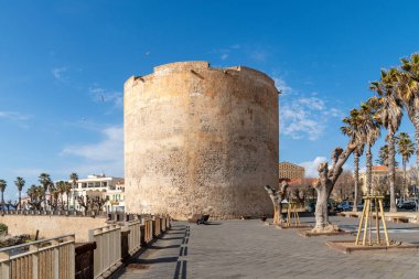 Alghero, Sardinia - İtalya - 02-13-2025: Kurumuş ağaçlar ve palmiyelerle çevrili, yıpranmış bir yüzeyi olan devasa yuvarlak bir taş kule. Modern binalar ve gökyüzü.