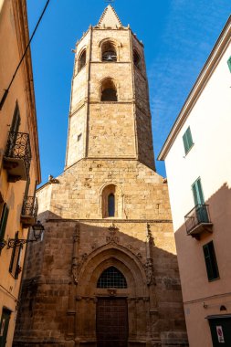 Alghero, Sardinia - İtalya - 02-13-2025: Gotik kemerli tarihi bir çan kulesi ve mavi gökyüzüne karşı sivri bir çatı yükseliyor. Balkonlu binalar dar sokağı çerçeveliyor..