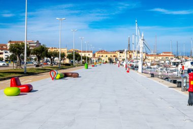 Palau - Sardinien - Italien - 02-19-2025: Modern renkli banklar, parlak mavi gökyüzü altında demirlemiş yelkenli tekneleri, binaları ve ağaçlarıyla Sardunya 'daki Palau Marina' nın gezinti alanı..