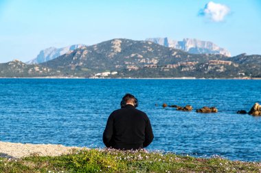 Olbia - Sardinien - Italien - 02-18-2025: Kulaklıklı bir adam uzak arkaplanda engebeli dağlarla Olbia yakınlarındaki sakin denize bakıyor.
