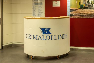 Barselona, Katalonya - İspanya - 01-10-2025: Tekerlekli Grimaldi Hatları logosu olan beyaz resepsiyon masası, kırmızı duvarlı bir feribot koridoruna yerleştirildi, tabelalar ve kaldırımlı bir caddenin fotoğrafı.