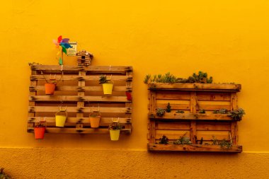 Alghero, Sardinia - İtalya - 02-13-2025: Parlak sarı bir duvardaki iki ahşap palet dikey bahçeler olarak hizmet veriyor. Renkli tencereler, sulandırıcılar ve bir yel değirmeni canlı bir görüntü oluşturur..