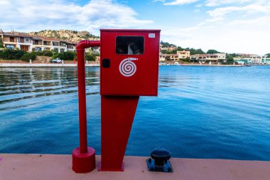 Porto Cervo - Sardinien - Italien - 02-22-2025: Porto Cervo, Sardinya 'nın rıhtım tarafında sakin rıhtım, meskun evler ve yukarıda mavi gökyüzü bulunan kırmızı bir itfaiye hortumu bulunuyor.