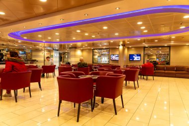 Barselona, Katalonya - İspanya - 01-10-2025: Kırmızı sandalyeli bir feribot salonu, bar alanı, oturan yolcular ve sıcak ışıklandırma ve mavi tavan ışıkları altında duvarda iki televizyon.