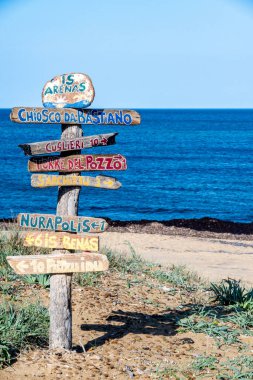 SArchittu - Sardinien - Italien - 03-02-2025: Sarkittu, Sardinya, İtalya 'da mavi deniz ve açık gökyüzü olan kumlu bir plajda renkli yön işaretleriyle ahşap tabela.