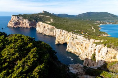 Alghero - Sardinien - Italien - 02-16-2025: Yukarıdaki yeşil ormanlık sarp kayalıklar ve aşağıdaki turkuaz deniz, mavi gökyüzü koşulları altında görünen kıvrımlı kıyı yolu.
