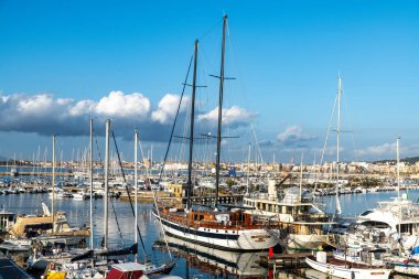 Alghero, Sardinia - İtalya - 02-13-2025: Rıhtıma demirlemiş yelkenli ve motorlu tekneleri olan bir marina. Siyah direkli ve tahta güverteli büyük bir yelkenli gemi. Su tekneleri ve gökyüzünü yansıtır..