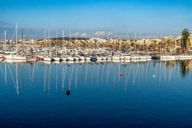 Alghero, Sardinia - İtalya - 02-13-2025: Yelkenliler sakin suya yansıyan uzun direklerle rıhtıma demirlediler. Parlak gökyüzü, kırmızı şamandıralar ve modern binalar karşıtlık yaratır..