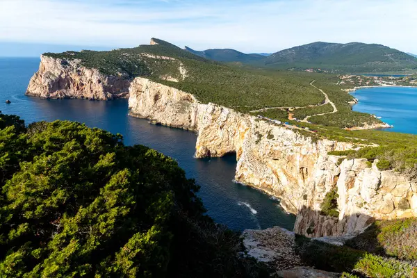 Alghero - Sardinien - Italien - 02-16-2025: Yukarıdaki yeşil ormanlık sarp kayalıklar ve aşağıdaki turkuaz deniz, mavi gökyüzü koşulları altında görünen kıvrımlı kıyı yolu.