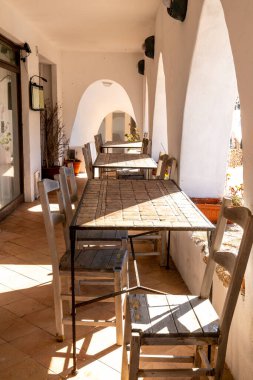 Porto Cervo - Sardinien - Italien - 02-22-2025: Porto Cervo, Sardunya 'da kırsal ahşap masalar ve beyaz kemerlerin altında sıcak güneş ışığıyla aydınlatılmış açık hava kafe koltukları.