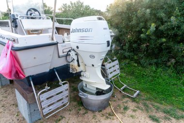 Nodu Pianu - Sardinien - Italien - 02-23-2025: Beyaz Johnson ek motoru katlanmış merdivenlerle küçük bir iskeleye monte edilmiş, otlar ve çalılarla çevrili kuru arazide dinleniyor.