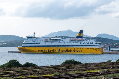 Golfo Aranci - Sardinien - Italien - 02-24-2025: Büyük Korsika Sardunya feribotu, arka planda sarı gövdesi, üst güverteleri ve dağlık kıyı şeridini gösteren bir limanda demirli..