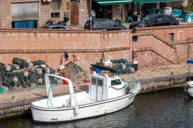 Bosa - Sardinien - Italien - 02-28-2025: Küçük beyaz bir balıkçı teknesi balıkçı ağları, halatları ve teçhizatları ile bir sokak kafesinin yakınındaki taş duvara kenetlenmiş..