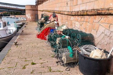 Bosa - Sardinien - Italien - 02-28-2025: Sayısız balıkçı tuzağı, halat, ağ ve teçhizat, suyun yanındaki taş rıhtım duvarı boyunca istiflenmiş, teknenin bir kısmı görülebiliyor.
