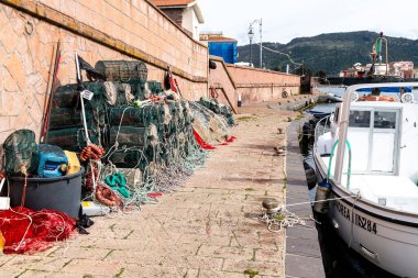 Bosa - Sardinien - Italien - 02-28-2025: Balıkçılık tuzakları, ağları ve ipleri, arka planda tepeler, lamba direkleri ve binaları bulunan demirlenmiş beyaz bir teknenin yanındaki taş iskele boyunca istiflenmiştir..