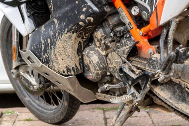 Bosa - Sardinien - Italien - 02-28-2025: Çamurlu bir macera motosiklet motoruna yakın plan, ayaklığı, vites değiştiricisini, motor kılıfını ve turuncu çerçeveyi fayanslı bir yüzeyde gösteriyor.