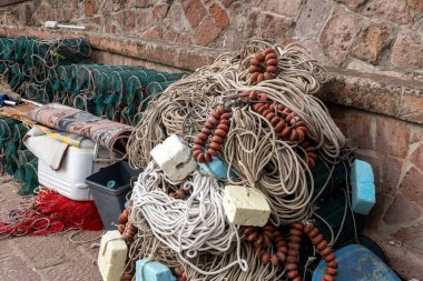 Bosa - Sardinien - Italien - 02-28-2025: Kahverengi yüzgeçli sıkıca sarmalanmış halatlar ve liman yolundaki taştan bir duvarın yanındaki yeşil tel balıkçı tuzaklarının yanındaki beyaz köpük şamandıraları.