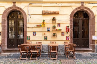 Bosa - Sardinien - Italien - 02-28-2025: Ahşap masa ve sandalyeler, çerçeve kapılar, duvar sanatı, saksı bitkileri, küçük raflar ve dekoratif sicim ışıklarıyla renkli bir bistro cephesi manzarası.