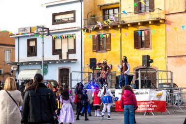 Bosa - Sardinien - Italien - 02-28-2025: Kostümlü çocuklar, renkli kiraz çiçekleriyle dekore edilmiş bir sokakta iki hippi kostümlü sanatçının eğlendirdiği bir sahnenin önünde duruyor.