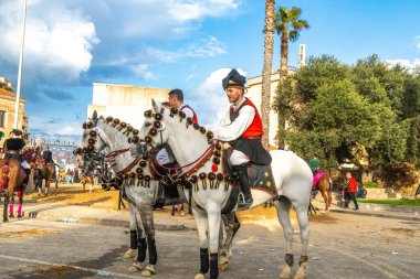 Oristano - Sardinien - Italien - 03-02-2025: Oristano karnavalında süslü beyaz atlara binen geleneksel kostümlü adamlar, palmiye ağaçları, mavi gökyüzü ve tarihi binalarla çevrili.