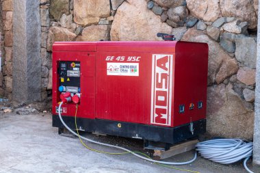 Porto Cervo - Sardinien - Italien - 02-22-2025: Sardinya 'nın Porto Cervo kentinde elektrik dağıtımı için kablolarla bağlı taşralı bir duvarın yanına kırmızı bir Mosa jeneratör birimi yerleştirildi.
