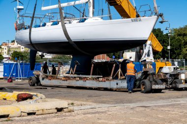 Cagliari - Sardinien - Italien - 03-10-2025: Yansıtıcı yelekli ve miğferli işçiler güneşli bir günde tersanede süreci izlerken bir yelkenli sarı bir vinç tarafından kaldırıldı.