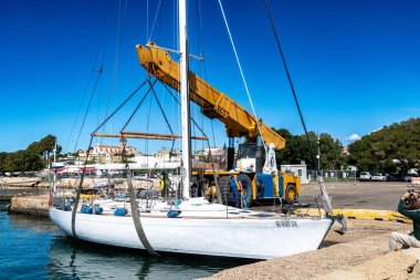 Cagliari - Sardinien - Italien - 03-10-2025: İşçiler açık mavi gökyüzü altında prosedürü denetlerken, iskelede sarı bir vinç tarafından sudan beyaz bir yelkenli kaldırılıyor.