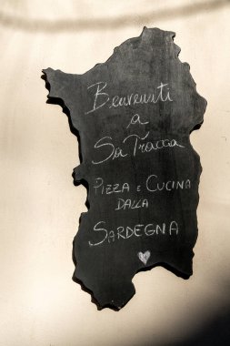 Cagliari - Sardinien - Italien - 03-11-2025: Sardinya şeklinde el yazısı tebeşirle yazılmış siyah bir tabela, Sa Tracca 'ya gelen misafirleri karşılayarak, adadan pizza ve mutfağı destekliyor..