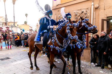 Oristano - Sardinien - Italien - 03-02-2025: Oristano Karnavalı sırasında süslemeli mavi kostümlü maskeli süvariler, kalabalık ve tarihi binalarla çevrili, şenlikli atlara biniyorlar.