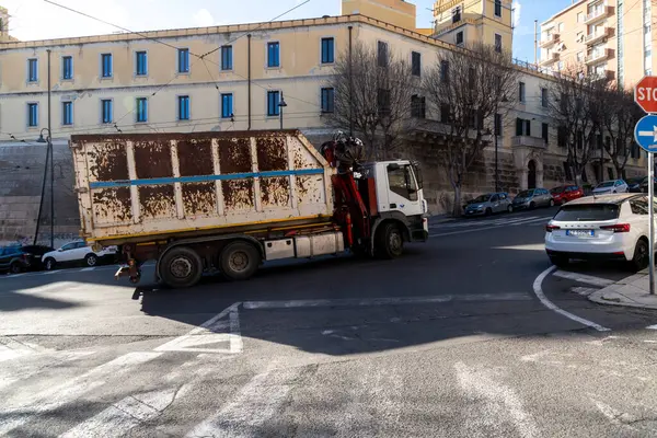 Cagliari - Sardinien - Italien - 03-11-2025: Vinçli paslı beyaz kamyon, park halindeki arabalar, binalar ve parlak güneş ışığı altında trafik işaretleriyle çevrili bir caddede dönüyor.