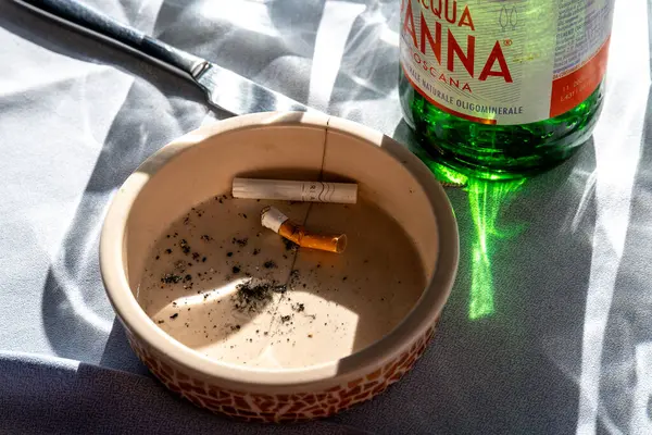 Cagliari - Sardinien - Italien - 03-11-2025: Açık havada güneş ışığıyla aydınlatılan yeşil cam su şişesi ve bıçağın yanında sigara izmaritleri ve külleri bulunan kül tablası.