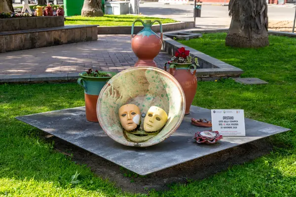 Oristano - Sardinien - Italien - 03-03-2025: Oristano 'da halka açık yeşil bir alanda sergilenen geleneksel terakotta kaplarıyla çevrili büyük bir seramik kasede iki tiyatro maskesi.