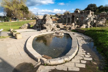 Fordongianus - Sardinien - Italien - 03-04-2025: Oval taş havzası Fordongianus, Sardinya 'daki Roma hamamlarında antik harabeler, kaldırımlar ve yeşilliklerle çevrili, parlak mavi bir gökyüzü altında.