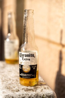 Fordongianus - Sardinien - Italien - 03-04-2025: Güneşin aydınlattığı bir taş yüzeyi üzerinde görünen yoğunlaşma ve arka planda ikinci bir bulanık şişe bulunan yarısı boş bir Coronita bira şişesi duruyor.