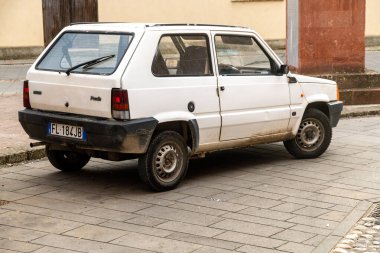 Fordongianus - Sardinien - Italien - 03-04-2025: Altında toprak olan beyaz bir Fiat Panda, sarı ve kırmızı dış duvarları olan taştan bir binanın yanındaki kaldırımda park edilmiştir..