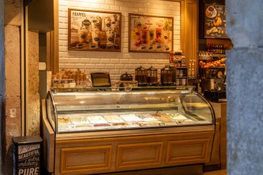 Reus, Katalonya - İspanya - 01-03-2025: Arka planda waffle konileri, içecek menüsü ve kahve yapma ekipmanları bulunan samimi bir dondurmacıda aydınlatılmış dondurma tezgahı.