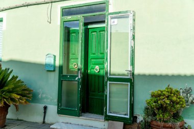 Semestene, Sardinya - İtalya - 01-12-2025: Semestene 'de cam yan panelleri olan yeşil ahşap bir kapı, aşınma belirtilerini gösteriyor, saksı bitkileriyle birlikte pastel yeşil bir duvara yaslanmış..