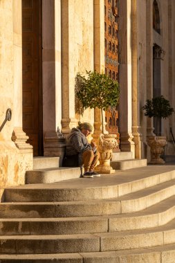 Cagliari - Sardinien - Italien - 03-11-2025: Bir kadın, süslü ahşap bir kapının önündeki altın güneş ışığıyla aydınlatılmış saksı ağacının yanındaki katedral basamaklarında oturuyor..