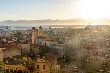 Cagliari - Sardinien - Italien - 03-11-2025: Kubbeli kiliseler, çatılar, yolcu gemileri ve liman manzaralı Cagliari şehri, yumuşak günbatımı ışığında dağlarla çevrili.