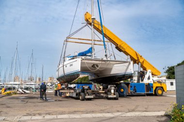Cagliari - Sardinien - Italien - 03-14-2025: Katamaran, arka planda rıhtımdaki işçiler ve limanlı yelkenlilerin direkleriyle çevrili bir marinadaki sarı bir vinç tarafından kaldırıldı.