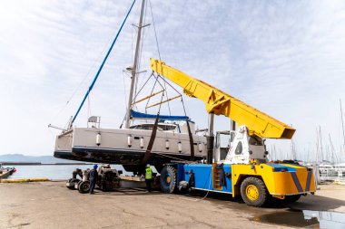 Cagliari - Sardinien - Italien - 03-14-2025: Bir katamaran sarı bir vinç ve tersanedeki işçiler tarafından kıyıya yanaşmış yelkenlilerin direkleriyle çevrili olarak sudan çıkarıldı.