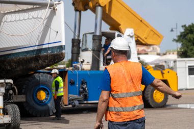 Cagliari - Sardinien - Italien - 03-14-2025: Turuncu yelek ve kask giyen bir ustabaşı, yoğun bir marinadaki vinçle bir katamaranı güpegündüz kaldıran bir takımı denetliyor.