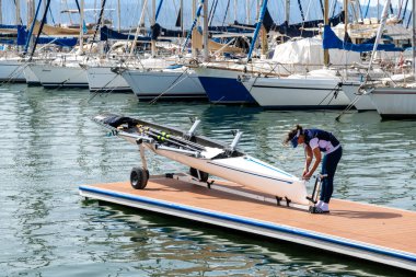 Cagliari - Sardinien - Italien - 03-14-2025: Bir dişi kürekçi, açık gökyüzünün altındaki bir marinada demirlemiş yelkenlilerle çevrili yüzen bir rıhtımda tek bir kayık hazırlıyor.