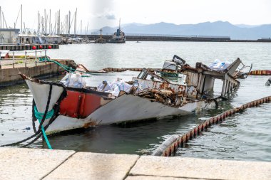Cagliari - Sardinien - Italien - 03-14-2025: Paslı, kısmen batık bir tekne, arka planda görünen diğer gemilerle birlikte yüzen bir bariyerle çevrili bir liman duvarının yanında yatıyor.
