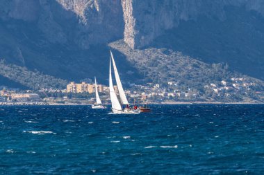 Palermo - Sizilien - Italien - 03-16-2025: Yükseltilmiş yelkenli iki beyaz yelkenli Palermo kıyısı yakınlarındaki dalgalı mavi sularda arka planda dağlarla yol alır.