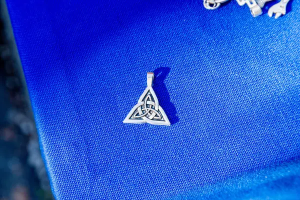 Jalon, Alicante - İspanya - 12-14-2024: Karmaşık düğüm tasarımlı gümüş bir Kelt Triquetra kolyesi, desenli mavi kumaş arka planda dinleniyor, ayrıntılı bir görüşe göre güneş ışığını yansıtıyor.