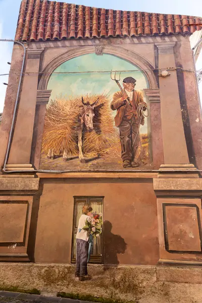 Semestene, Sardinya - İtalya - 01-12-2025: Kırsal bir binadaki bir duvar resmi, bir yaba ve bir eşekle saman taşıyan, bir kemer çerçeveli, aşağıda boyalı bir figürle bir çiftçiyi gösteriyor.